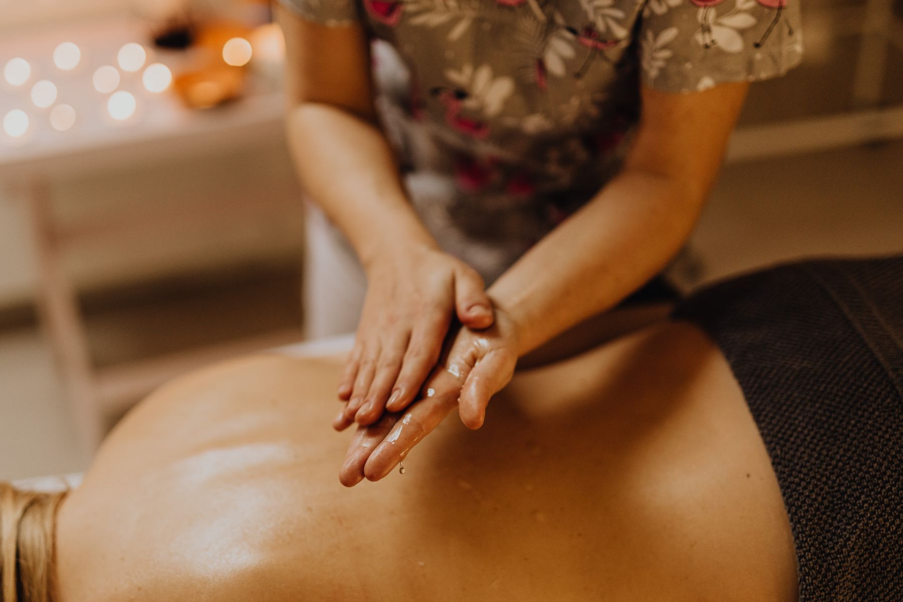 Mains d'une praticienne préparant une huile de massage au-dessus du dos d'une cliente dans une lumière chaude.
