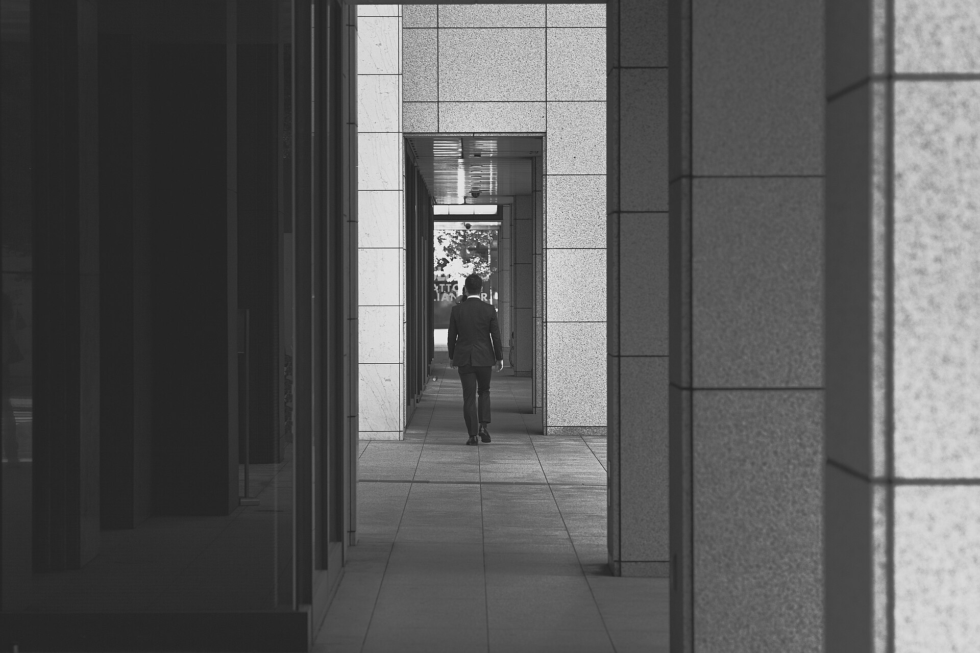 Galerie extérieure d’un quartier d’affaires, en noir et blanc : piliers de pierre alignés en enfilade et silhouette lointaine, de dos.