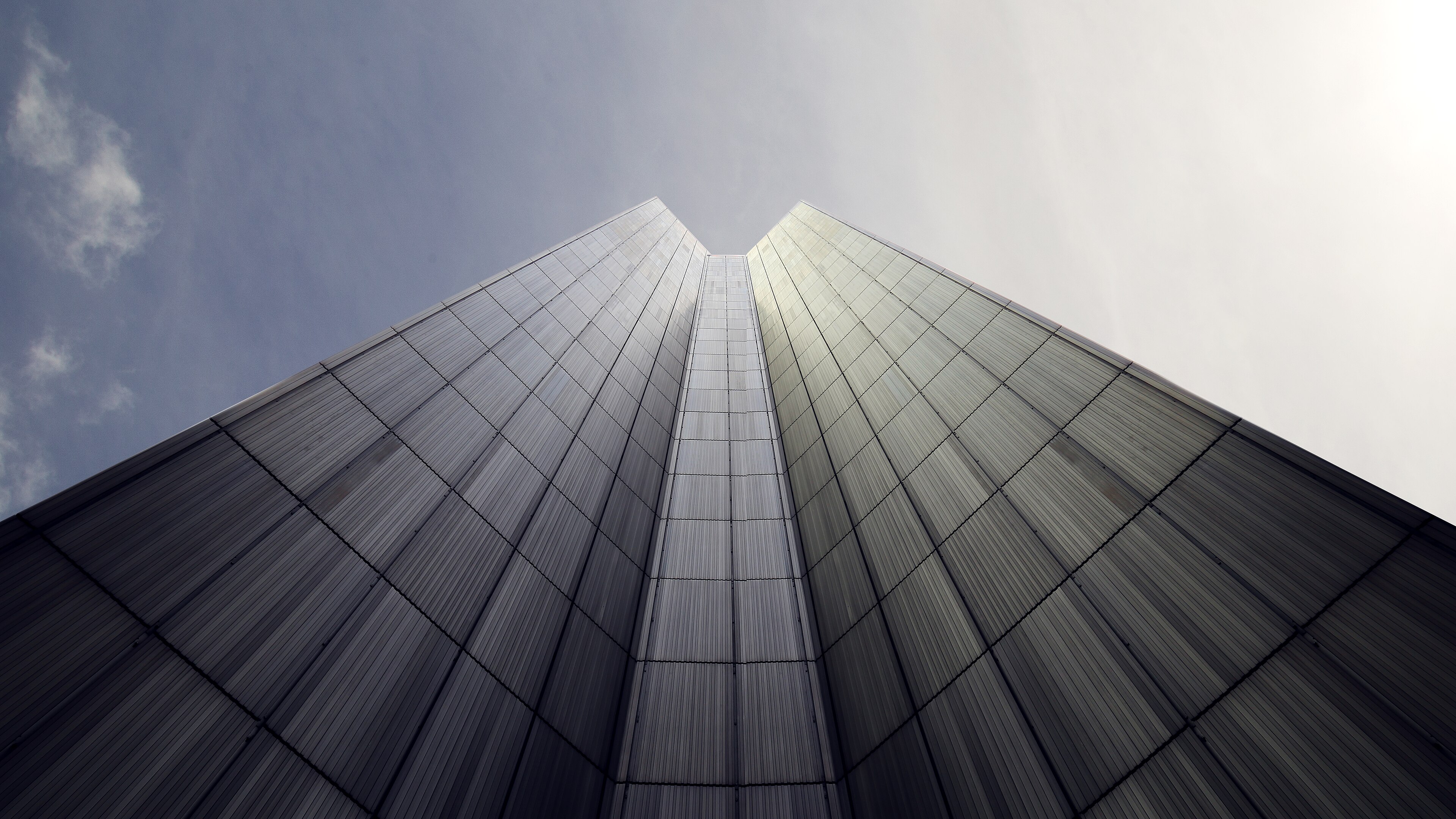 Tour de bureaux photographiée depuis son pied : deux façades vitrées convergent en une arête verticale qui file vers le ciel.