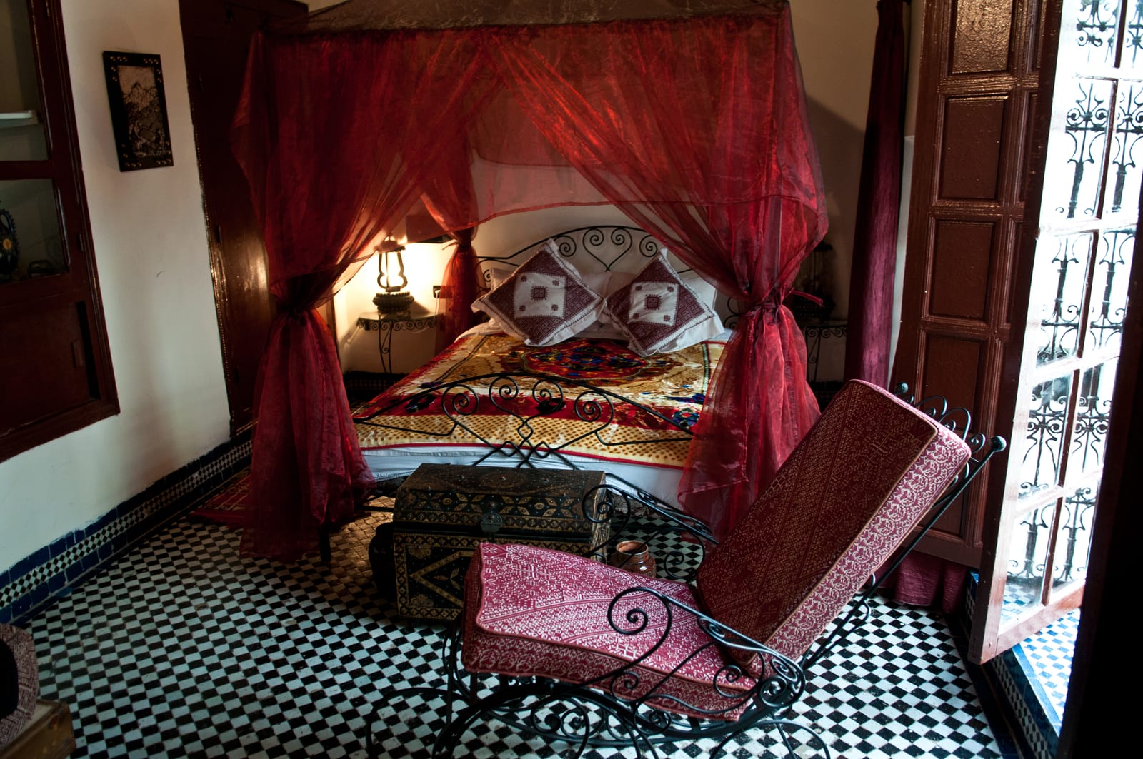 Chambre de riad avec lit à baldaquin drapé de rouge, zellige noir et blanc, mobilier en fer forgé et portes de bois ouvertes sur la lumière.