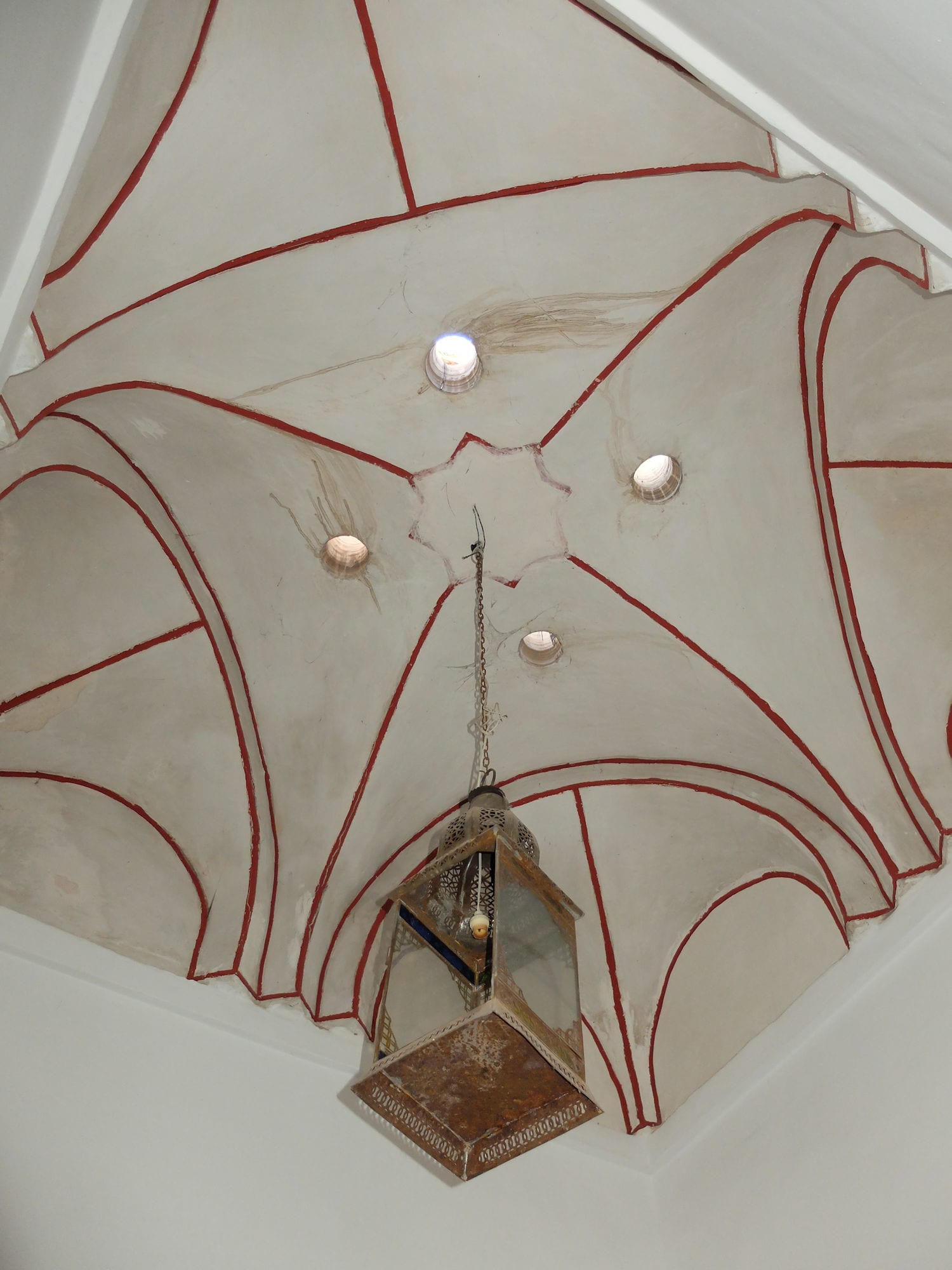 Voûte claire d'un hammam de Marrakech, soulignée de lignes rouges, percée d'oculi et éclairée par une lanterne en métal.