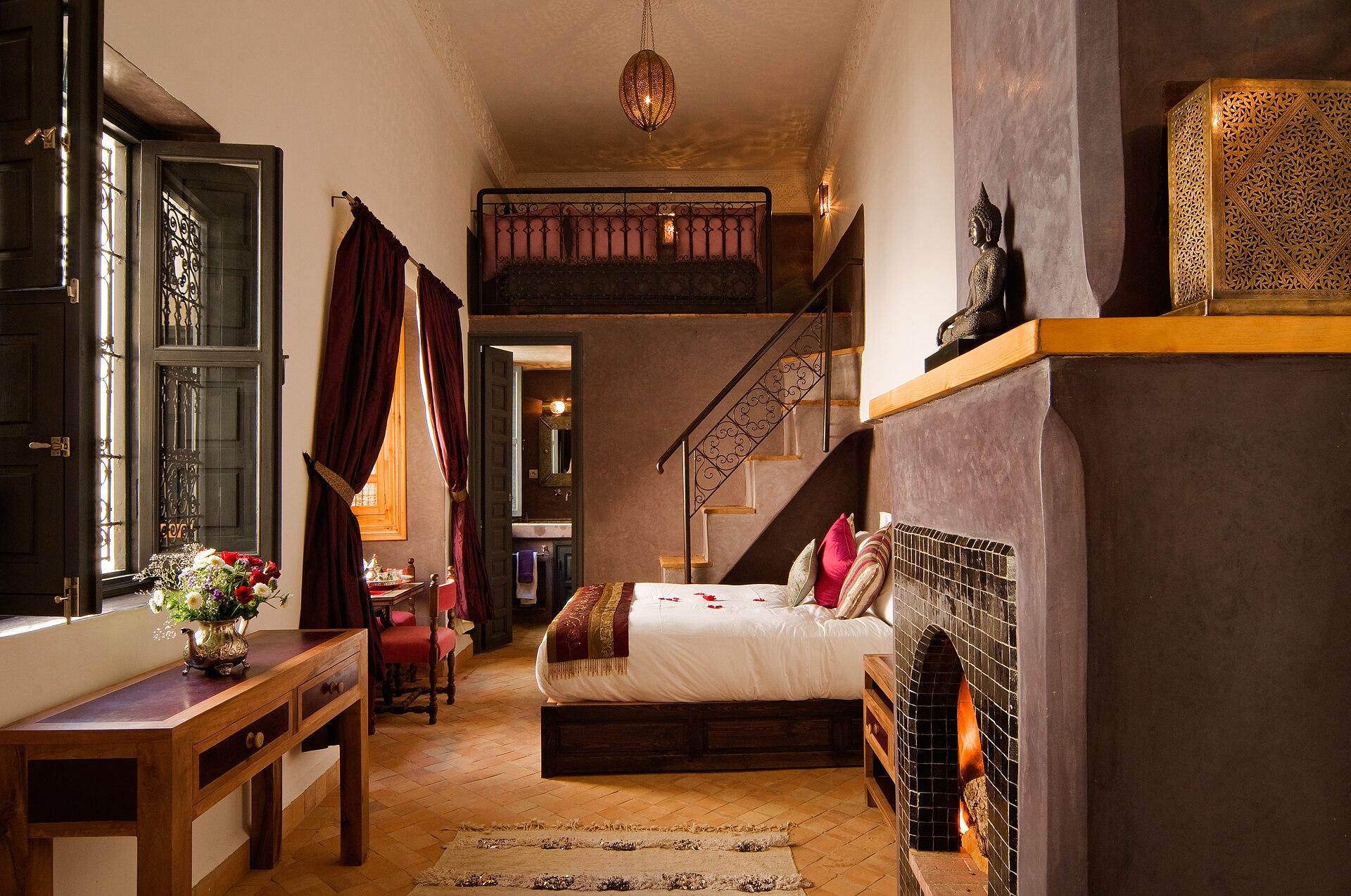 Suite sous haut plafond : cheminée allumée habillée de zellige, mezzanine de bois, escalier étroit, lit bas et volets de bois ouverts sur la lumière du jour.
