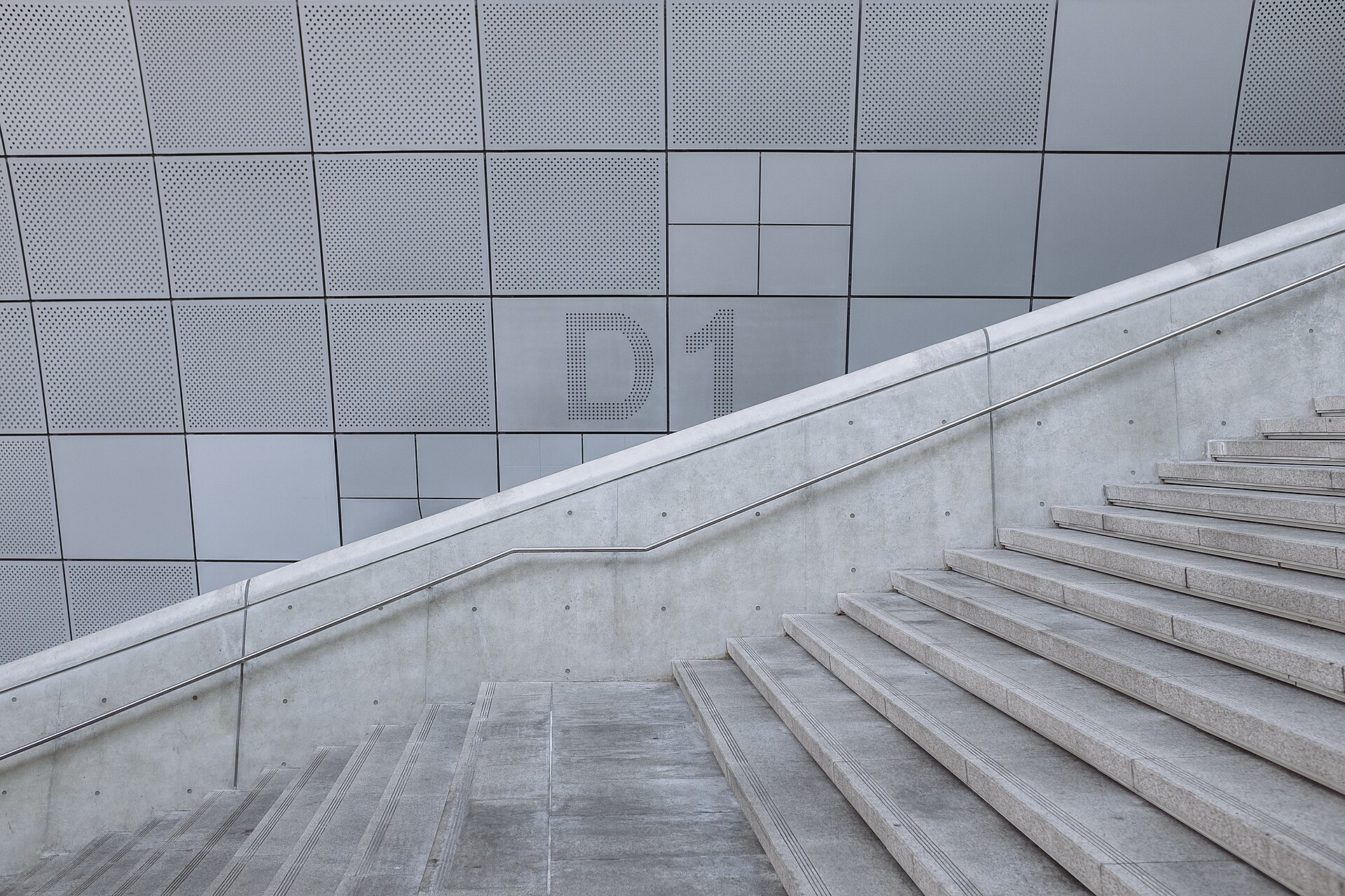 Escalier extérieur en béton bordé d'une main courante, longeant un mur de panneaux métalliques perforés.