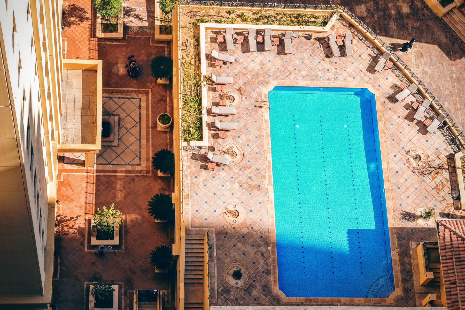 Vue aérienne d'une piscine bordée de chaises longues, eau turquoise et dalles claires.