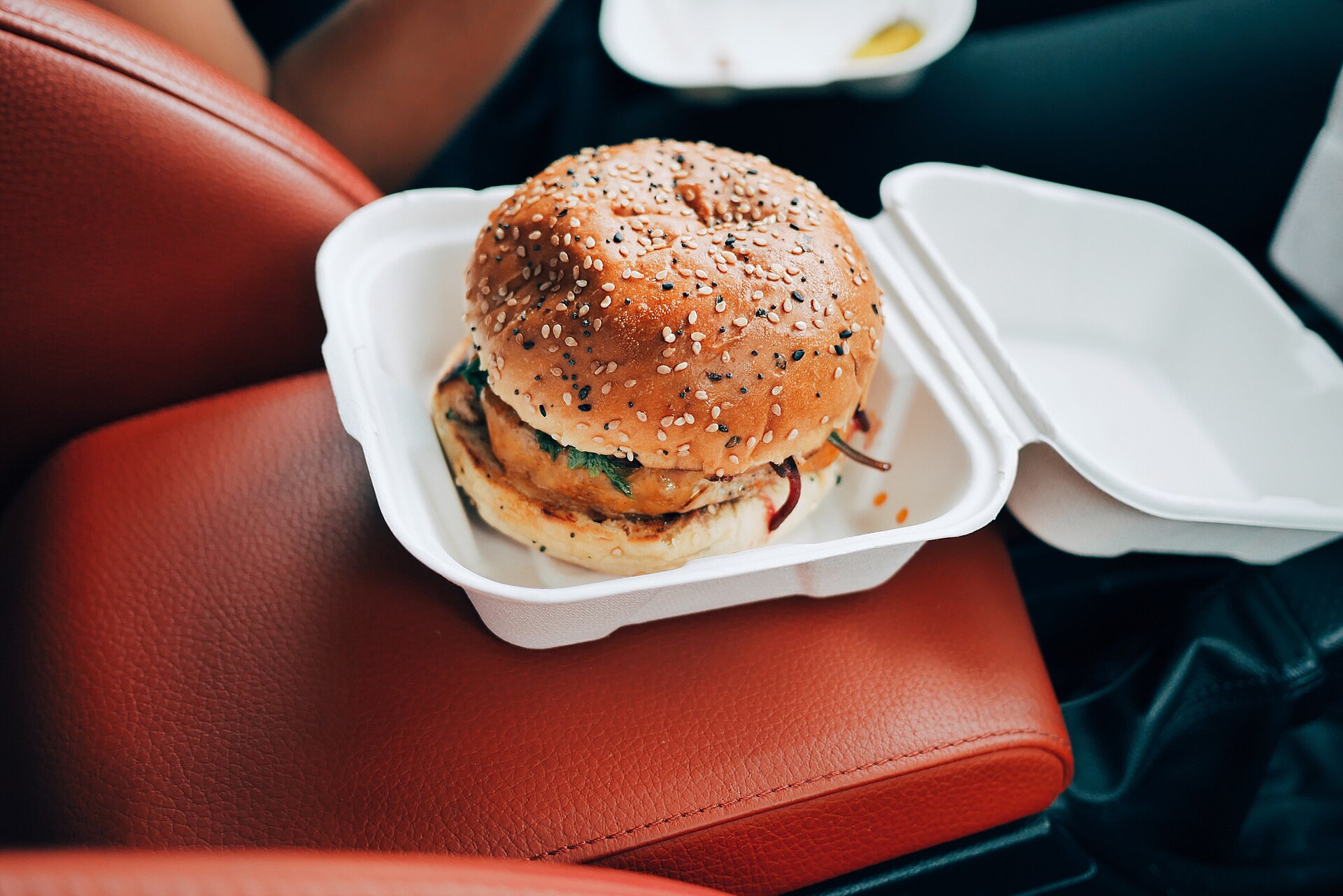 Burger au pain de sésame dans sa boîte à emporter ouverte, posée sur une banquette en cuir rouge.