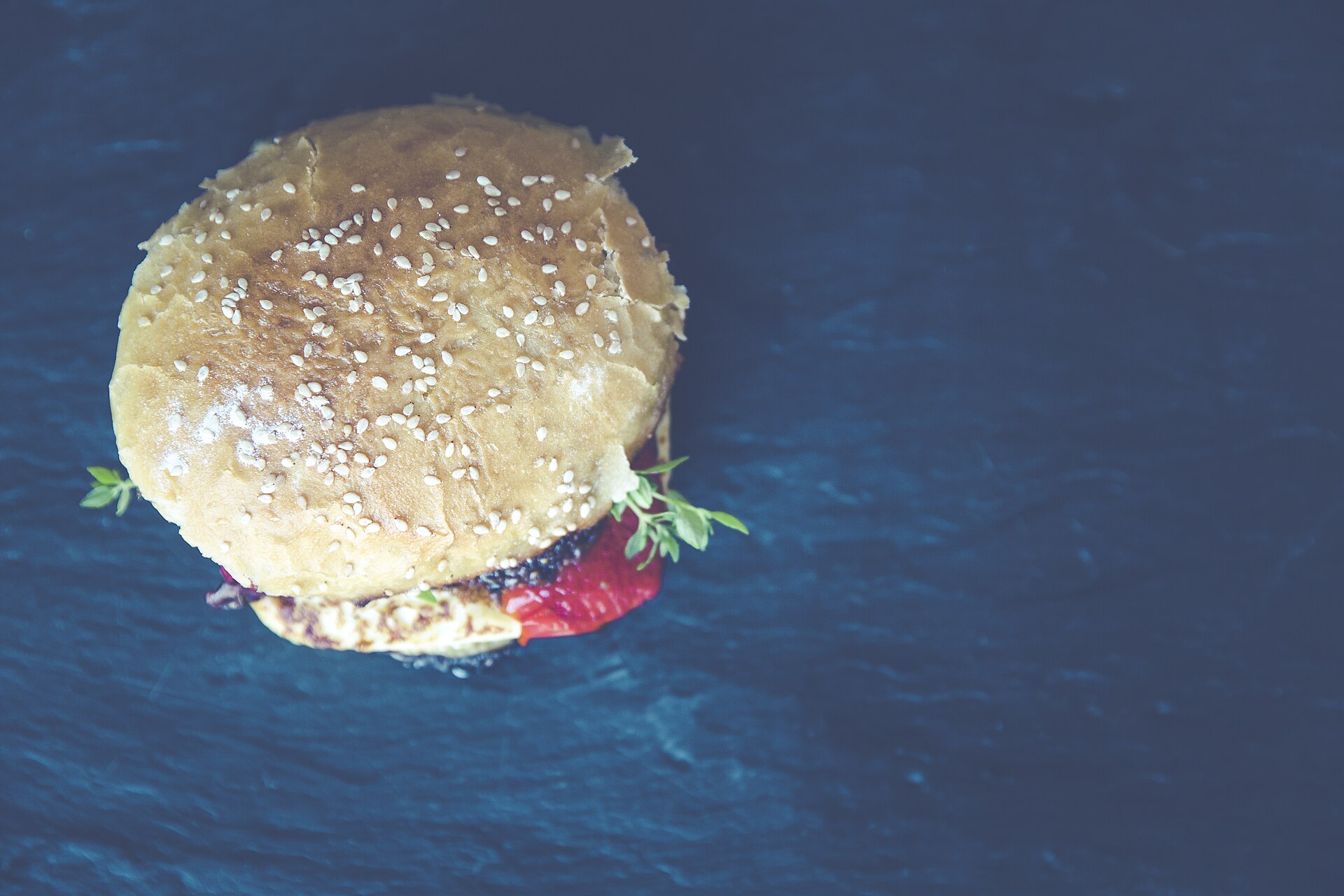 Vue de dessus d'un burger au pain de sésame posé sur une large ardoise noire, largement dégagée sur la droite.