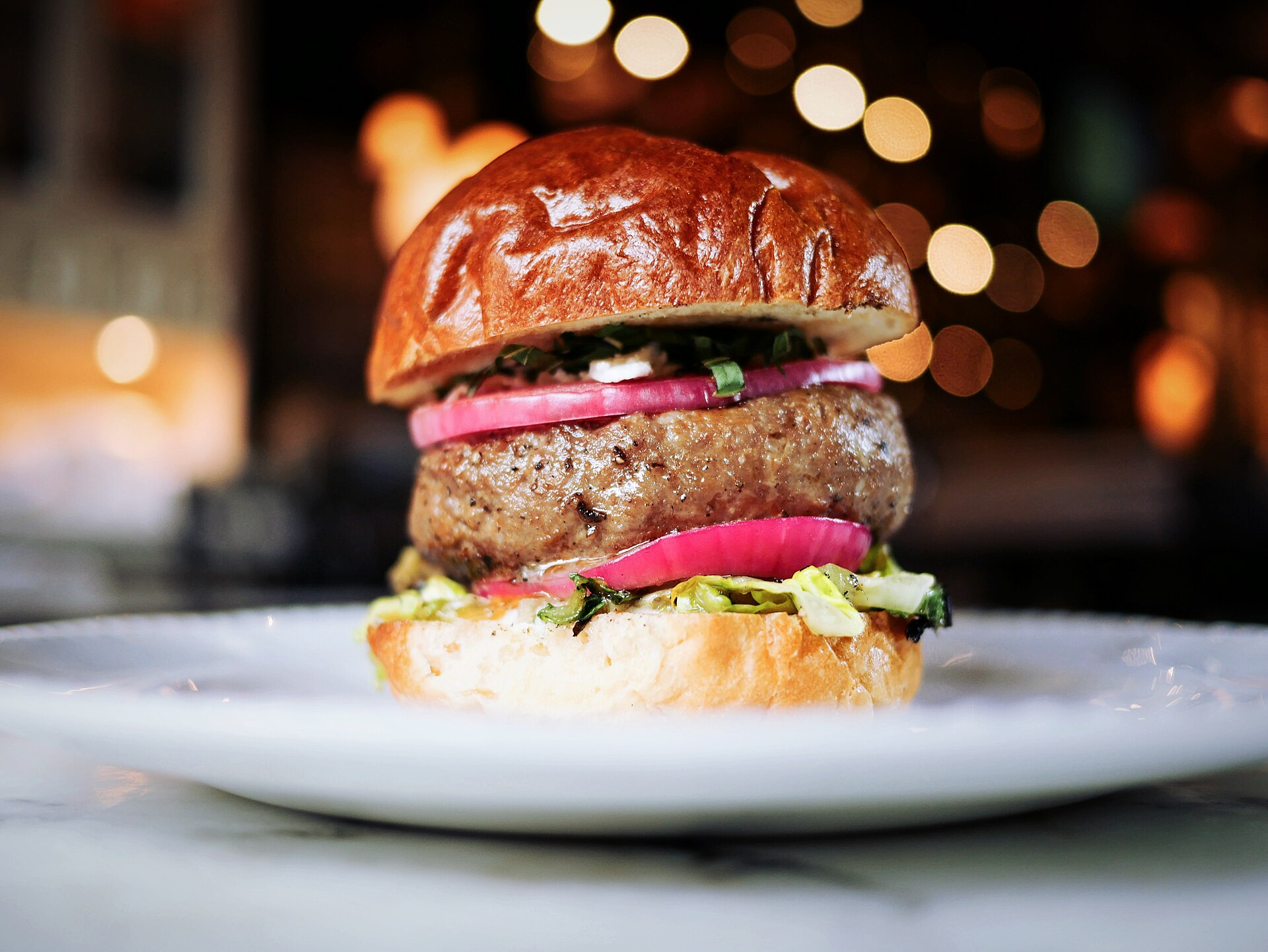 Burger au pain brioché doré photographié de profil sur une assiette claire, devant un fond sombre piqué de lumières chaudes floues.