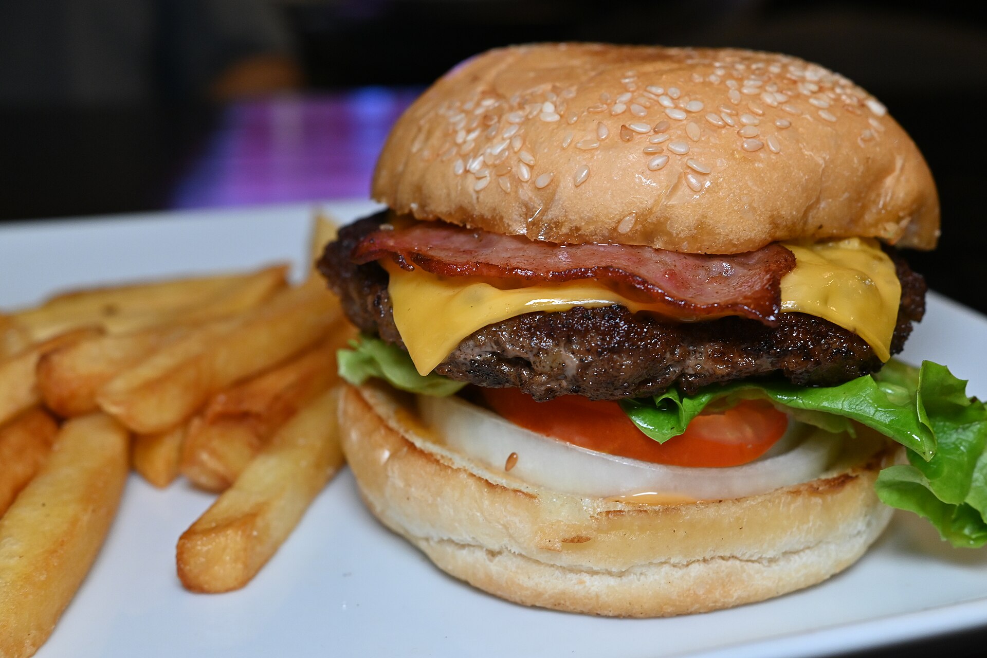 Cheeseburger au bacon et aux crudités servi avec une poignée de frites dorées sur une assiette blanche, fond sombre.