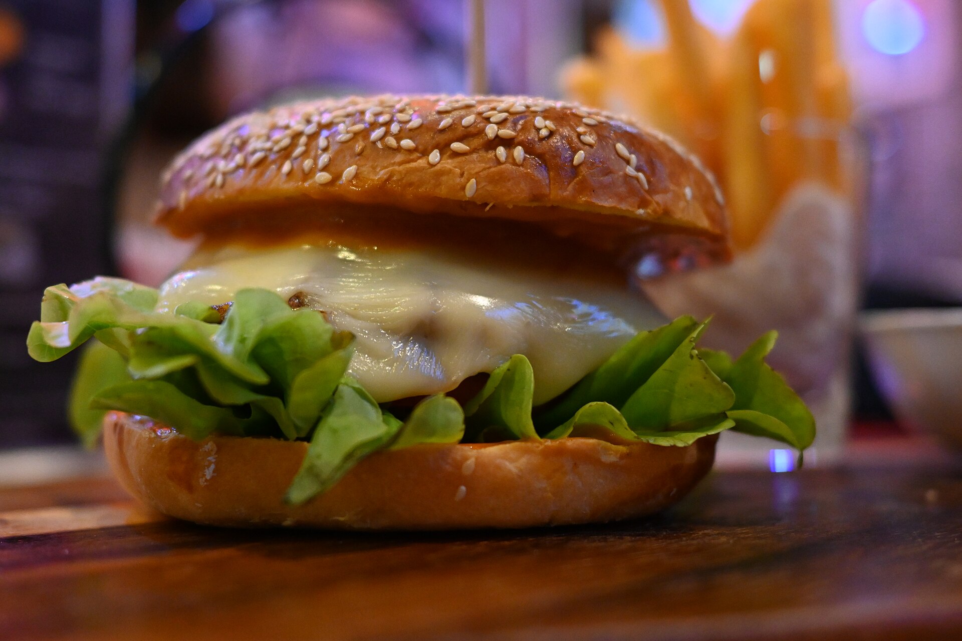 Macro d'un burger au pain de sésame : fromage fondu qui coule sur la salade, cornet de frites flou en arrière-plan, reflets colorés.