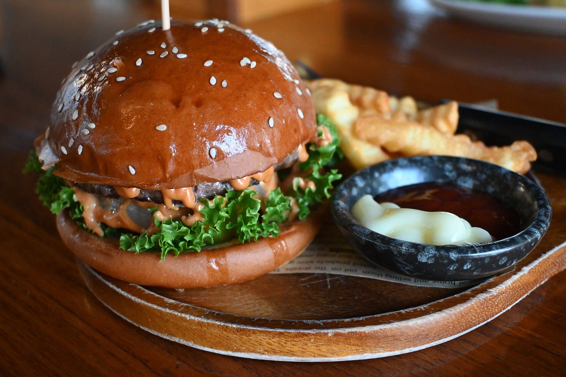 Burger au pain de sésame brillant servi sur une planche en bois avec des frites et deux sauces en coupelle.
