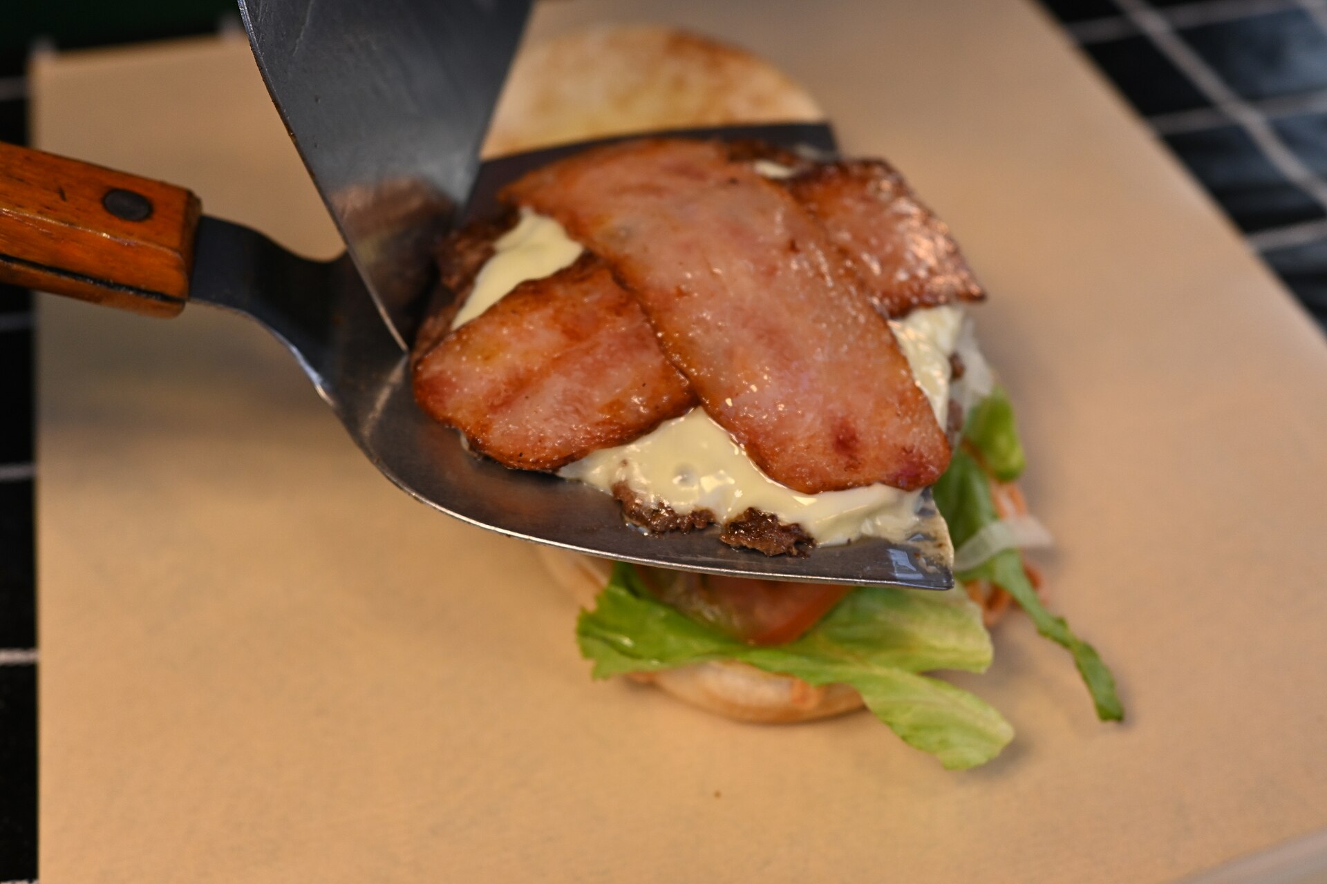 Tranches de bacon grillé déposées à la spatule sur un steak nappé de fromage fondu, juste avant la pose du pain.