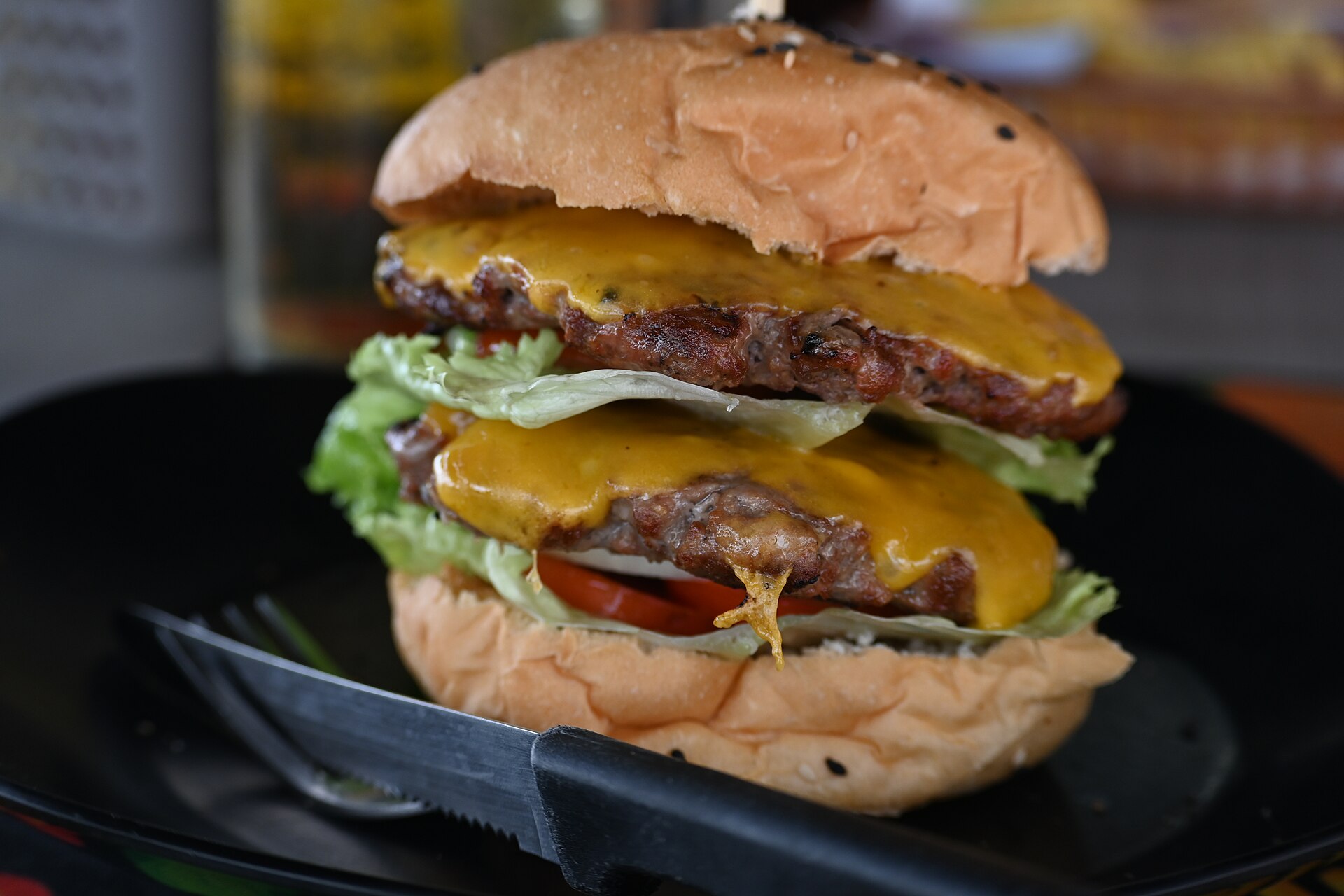 Gros plan d'un double cheeseburger : deux steaks épais nappés de cheddar fondu, salade craquante et tomate, entre deux pains briochés, sur une assiette noire mate.