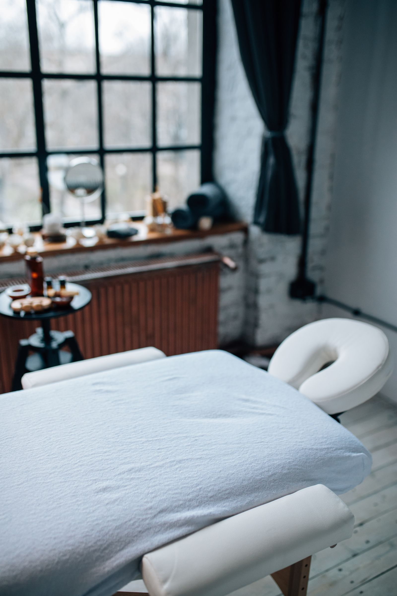 Cabine de soin avec table de massage blanche, linge roulé et grande fenêtre en lumière naturelle.