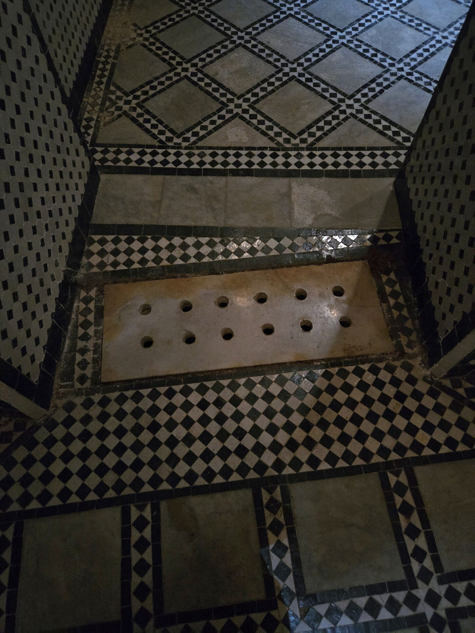 Détail d'un sol de bain traditionnel : dalle de marbre percée de douze trous pour laisser monter la vapeur, encadrée de mosaïques noires et blanches en damier.